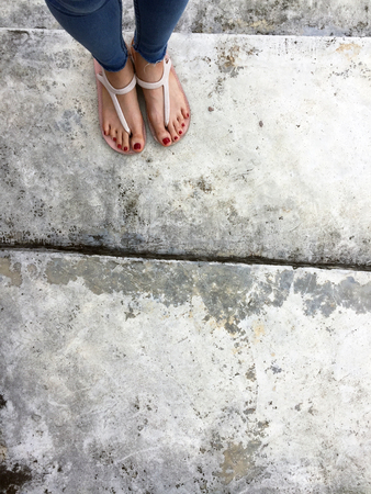 Close up of Bare Feet with Red Nail in Sandals and Blue Jeans Woman On The Concrete Floor Background Great For Any Use.の写真素材