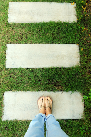Womanâs Legs and Feet Wear Gold Sandals on Cement and Green Grass Background Great for Any Use.の写真素材