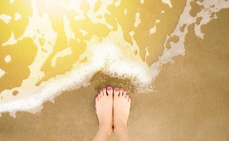 Selfie feet on beach background. Woman leg and foot with pink pedicure in summer. Top view of the barefoot in sand with wave motion coming to the feet - foaming sea. Nature textures concept.の写真素材