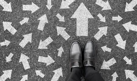 Feet and arrows on road background from above. Businessman standing on pathway with drawn white many direction arrows choice. Top view of A business man black shoes. Motivation and growth concept.の写真素材