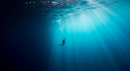 A lone diver descends into the deep blue ocean, illuminated by sun rays piercing through the waters surface, creating a serene underwater scene.の素材