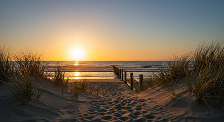 A breathtaking sunset over a sandy beach, with a wooden walkway leading to the ocean, framed by dune grass under a vibrant sky.の素材