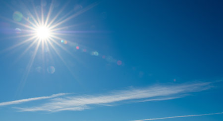 A radiant sun dominates a clear blue sky, accompanied by delicate wispy clouds and contrails, creating a serene and picturesque scene.の素材