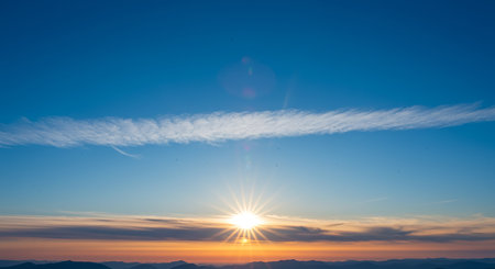 A vibrant sunrise paints the sky with orange and yellow hues, contrasting against the clear blue sky and wispy clouds above the horizon.の素材