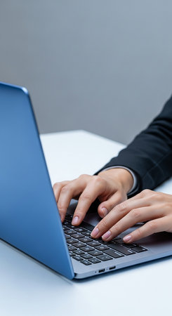 A close up vertical shot of a person in a dark suit typing on a blue laptop. The scene is set in a minimalist office environment with a white desk and grey background.の素材