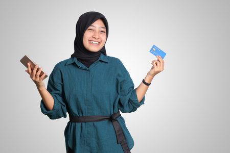 Portrait of cheerful Asian muslim woman with hijab, showing credit card while holding mobile phone. Financial and savings concept. Isolated image on white backgroundの写真素材