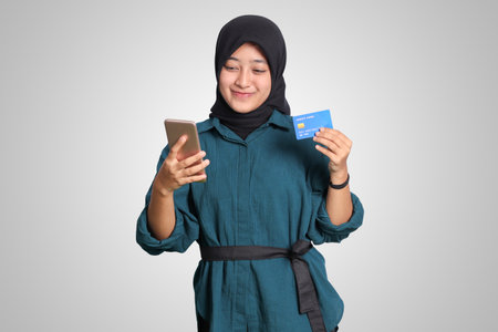 Portrait of cheerful Asian muslim woman with hijab, showing credit card while holding mobile phone. Financial and savings concept. Isolated image on white backgroundの写真素材