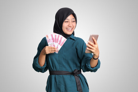 Portrait of excited Asian hijab woman in casual outfit showing one hundred thousand rupiah while holding a mobile phone. Financial and savings concept. Isolated image on white backgroundの写真素材