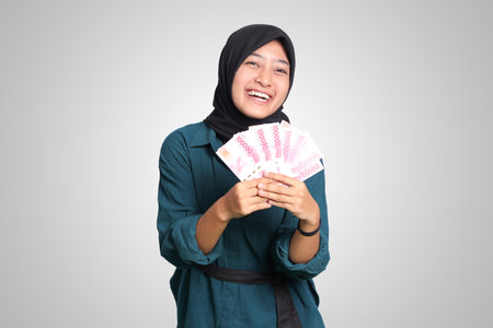 Portrait of excited Asian hijab woman in casual outfit showing one hundred thousand rupiah. Financial and savings concept. Isolated image on white backgroundの写真素材