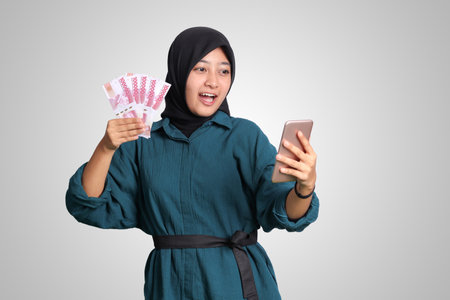 Portrait of excited Asian hijab woman in casual outfit showing one hundred thousand rupiah while holding a mobile phone. Financial and savings concept. Isolated image on white backgroundの写真素材