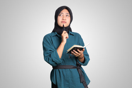 Portrait of excited Asian muslim woman with hijab writing on note book and thinking with pen on chin. Advertising concept. Isolated image on white backgroundの写真素材
