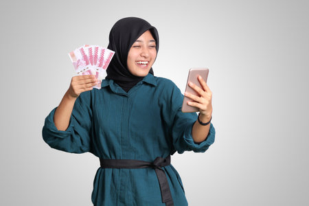 Portrait of excited Asian hijab woman in casual outfit showing one hundred thousand rupiah while holding a mobile phone. Financial and savings concept. Isolated image on white backgroundの写真素材