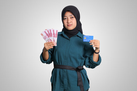 Portrait of cheerful Asian muslim woman with hijab, showing one hundred thousand rupiah while holding a credit card. Financial and savings concept. Isolated image on white backgroundの写真素材