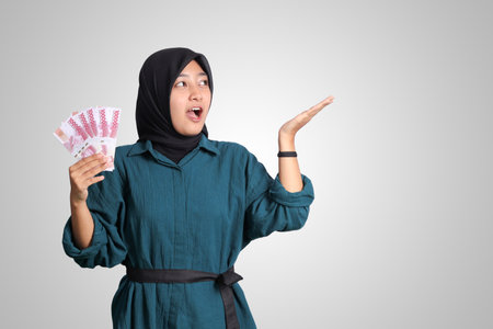 Portrait of excited Asian hijab woman in casual outfit showing and pointing one hundred thousand rupiah. Financial and savings concept. Isolated image on white backgroundの写真素材