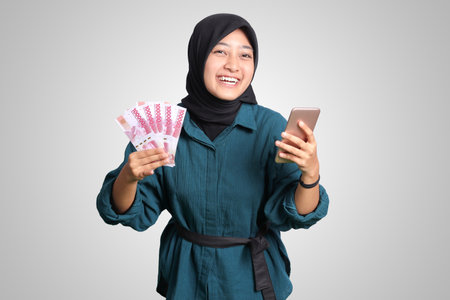 Portrait of excited Asian hijab woman in casual outfit showing one hundred thousand rupiah while holding a mobile phone. Financial and savings concept. Isolated image on white backgroundの写真素材