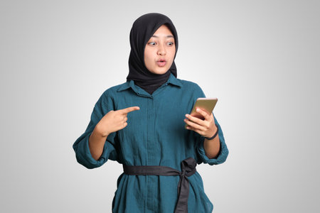 Portrait of excited Asian muslim woman with hijab holding and touching mobile phone while pointing with finger. Advertising concept. Isolated image on white backgroundの写真素材