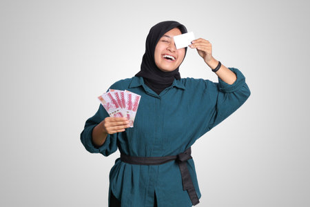 Portrait of cheerful Asian muslim woman with hijab, showing one hundred thousand rupiah while holding a credit card. Financial and savings concept. Isolated image on white backgroundの写真素材