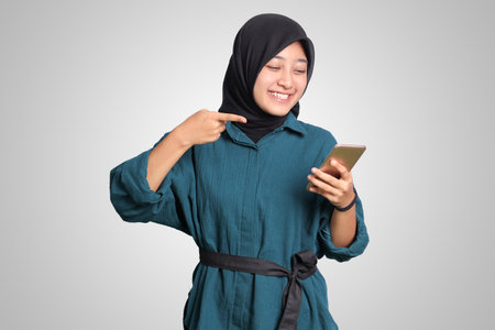 Portrait of excited Asian muslim woman with hijab holding and touching mobile phone while pointing with finger. Advertising concept. Isolated image on white backgroundの写真素材