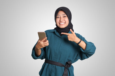 Portrait of excited Asian muslim woman with hijab holding and touching mobile phone while pointing with finger. Advertising concept. Isolated image on white backgroundの写真素材