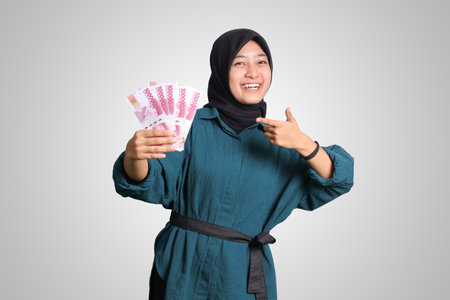 Portrait of excited Asian hijab woman in casual outfit showing and pointing one hundred thousand rupiah. Financial and savings concept. Isolated image on white backgroundの写真素材