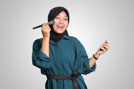 Portrait of excited Asian muslim woman with hijab, applying foundation powder or blush with makeup brush on her cheek. Isolated image on white backgroundの写真素材