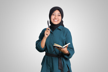 Portrait of excited Asian muslim woman with hijab writing on note book and pointing up with pen. Advertising concept. Isolated image on white backgroundの写真素材
