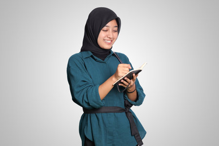 Portrait of excited Asian muslim woman with hijab writing on pocket note book with pen. Studying and learning concept. Isolated image on white backgroundの写真素材