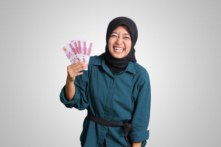 Portrait of excited Asian hijab woman in casual outfit showing one hundred thousand rupiah. Financial and savings concept. Isolated image on white backgroundの写真素材