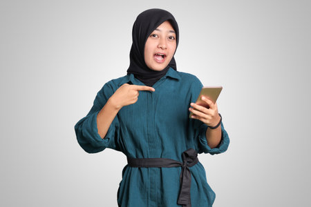 Portrait of excited Asian muslim woman with hijab holding and touching mobile phone while pointing with finger. Advertising concept. Isolated image on white backgroundの写真素材