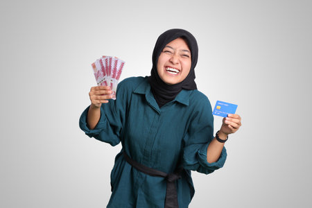 Portrait of cheerful Asian muslim woman with hijab, showing one hundred thousand rupiah while holding a credit card. Financial and savings concept. Isolated image on white backgroundの写真素材