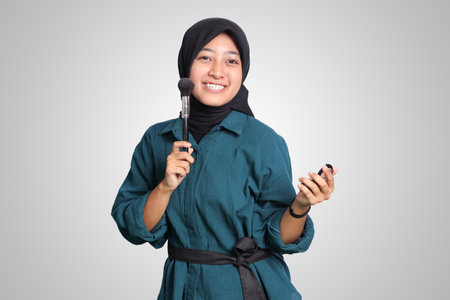 Portrait of excited Asian muslim woman with hijab, applying foundation powder or blush with makeup brush on her cheek. Isolated image on white backgroundの写真素材