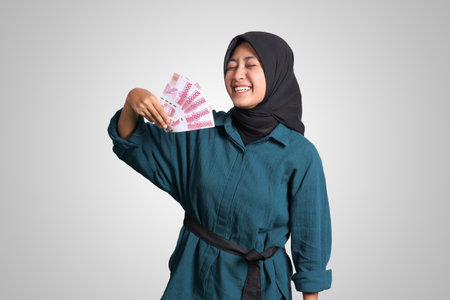 Portrait of excited Asian hijab woman in casual outfit showing one hundred thousand rupiah. Financial and savings concept. Isolated image on white backgroundの写真素材