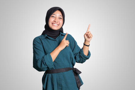 Portrait of excited Asian hijab woman in casual outfit standing against white background, showing product and pointing with hand and finger to the side. Advertising concept.の写真素材