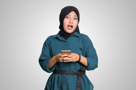 Portrait of shocked Asian muslim woman with hijab looking at her mobile phone. Wow face expression. Advertising concept. Isolated image on white backgroundの写真素材
