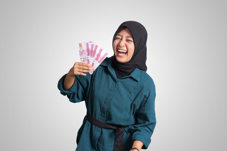 Portrait of excited Asian hijab woman in casual outfit showing one hundred thousand rupiah. Financial and savings concept. Isolated image on white backgroundの写真素材