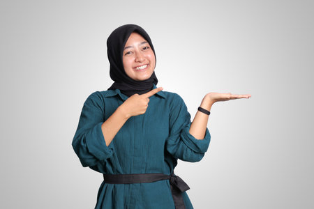 Portrait of excited Asian hijab woman in casual outfit standing against white background, showing product and pointing with hand and finger to the side. Advertising concept.の写真素材