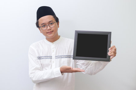 Portrait of young Asian muslim man holding a blackboard with hand pointed. Blank space for typography or text to fill. Isolated image on white backgroundの写真素材