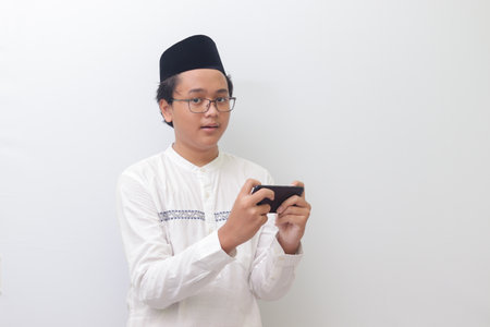 Portrait of young Asian muslim man raising his fist, celebrating winning game or getting good news on his mobile phone. Isolated image on white backgroundの写真素材