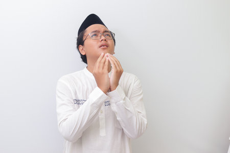Portrait of young millennial Asian muslim man praying earnestly with his hands raised. Isolated image on white backgroundの写真素材