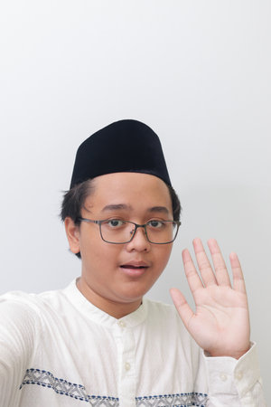 Portrait of smiling young Asian muslim man taking picture of himself or selfie, saying hi and waving his hand. Screen capture of a phone. Isolated image on white backgroundの写真素材