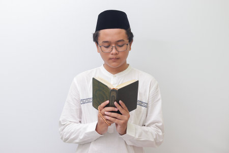 Portrait of young Asian muslim man reading and reciting Holy book of Quran seriously. Isolated image on white backgroundの写真素材