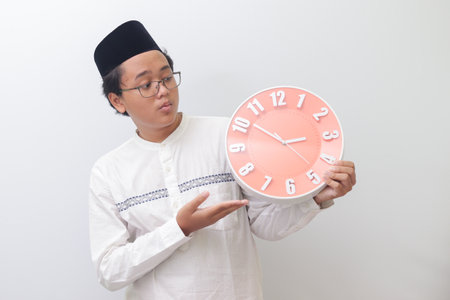 Portrait of young attractive Asian muslim man pointing and holding a clock that shows 3 am. Isolated image on white backgroundの写真素材