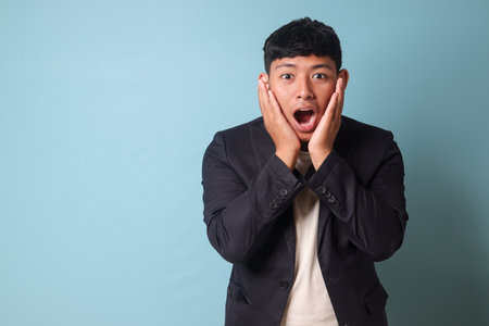 Portrait of young Asian business man in casualÂ suit surprised looking at camera with both hands in cheeks. Isolated image onÂ blueÂ backgroundの写真素材