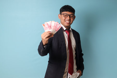 Portrait of young Asian business man in casualÂ suit smiling while holding thousand rupiahs proudly. Isolated image onÂ blueÂ backgroundの写真素材