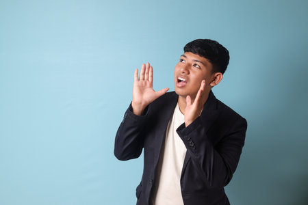 Portrait of young Asian business man in casualÂ suit shouting to the distance. Isolated image onÂ blueÂ backgroundの写真素材