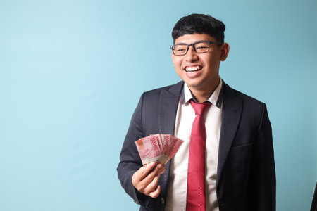 Portrait of young Asian business man in casualÂ suit laughing out loud holding thousands rupiahs. Isolated image onÂ blueÂ backgroundの写真素材