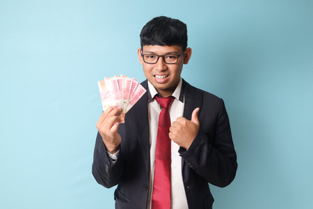 Portrait of happy young Asian business man in casual suit holding thousand rupiahs while making thumbs up. Isolated image on blue backgroundの写真素材