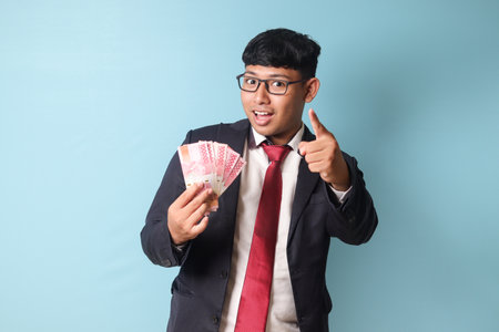 Portrait of young Asian business man in casualÂ suit pointing hand at camera while holding thousand rupiahs with persuasive expressions. Isolated image onÂ blueÂ backgroundの写真素材