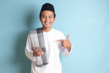 Portrait of young Asian muslim man holding mobile phone with smiling expression on face while pointing finger to the side. Isolated image on blue backgroundの写真素材