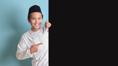 Portrait of young Asian muslim man showing product and pointing finger at copy space on big black paper poster advertising your text. Isolated image on blue backgroundの写真素材